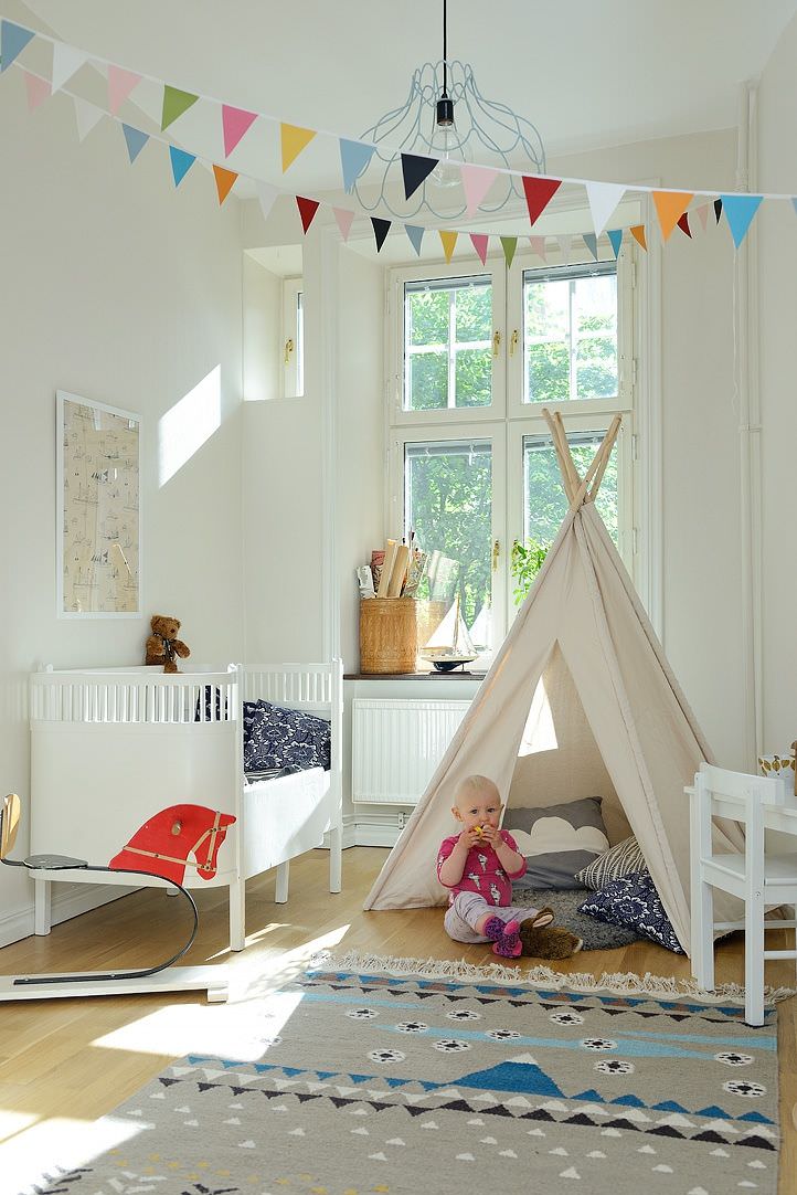 اتاق نوزاد با تخت سفید، کفپوش چوبی و چادر بازی که برای تزیین سقف آن از ریسه های کاغذی رنگارنگ استفاده شده است